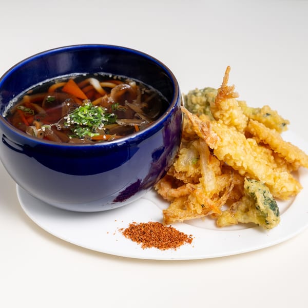 A blue bowl of Japanese udon noodle soup with tempura shrimp and vegetables on a white plate.